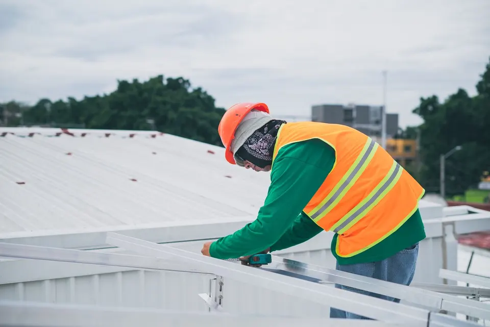 Worker in a hard hat and high-visibility vest measuring metal framing on a commercial rooftop